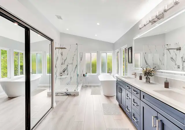 Laguna Hills primary bathroom with navy double vanity, quartz tops, freestanding soaking tub and marble-look walk-in shower with frameless glass