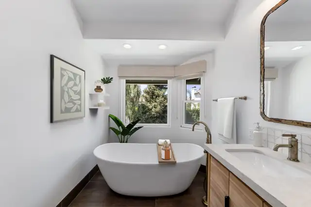 Santa Ana primary bathroom remodel with a freestanding white tub under a window and a wood vanity with brass fixtures